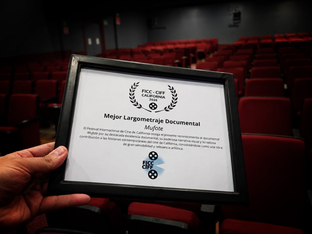 Certificado de reconocimiento a Mufote como Mejor Largometraje Documental en el Festival Internacional de Cine de California 2026, sostenido en una sala de cine con butacas rojas desenfocadas al fondo.