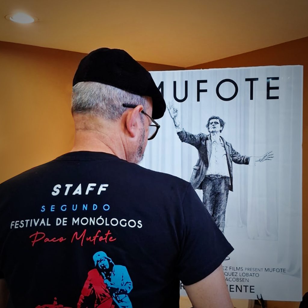 Salvador Pelayo-Frias con la playera del Festival de Monólogos Paco Mufote frente al cartel de MUFOTE